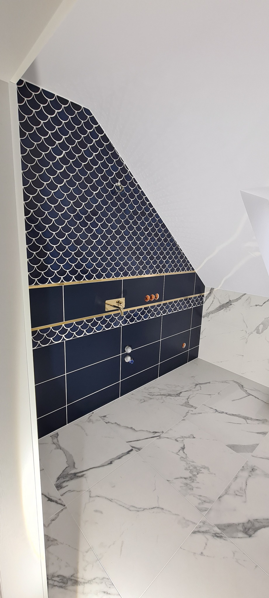 Wykończona łazienka na poddaszu z ciemnoniebieskimi płytkami w kształcie rybiej łuski na skośnej ścianie, złotymi akcentami i marmurową podłogą, widoczne podłączenia instalacji wodnej.