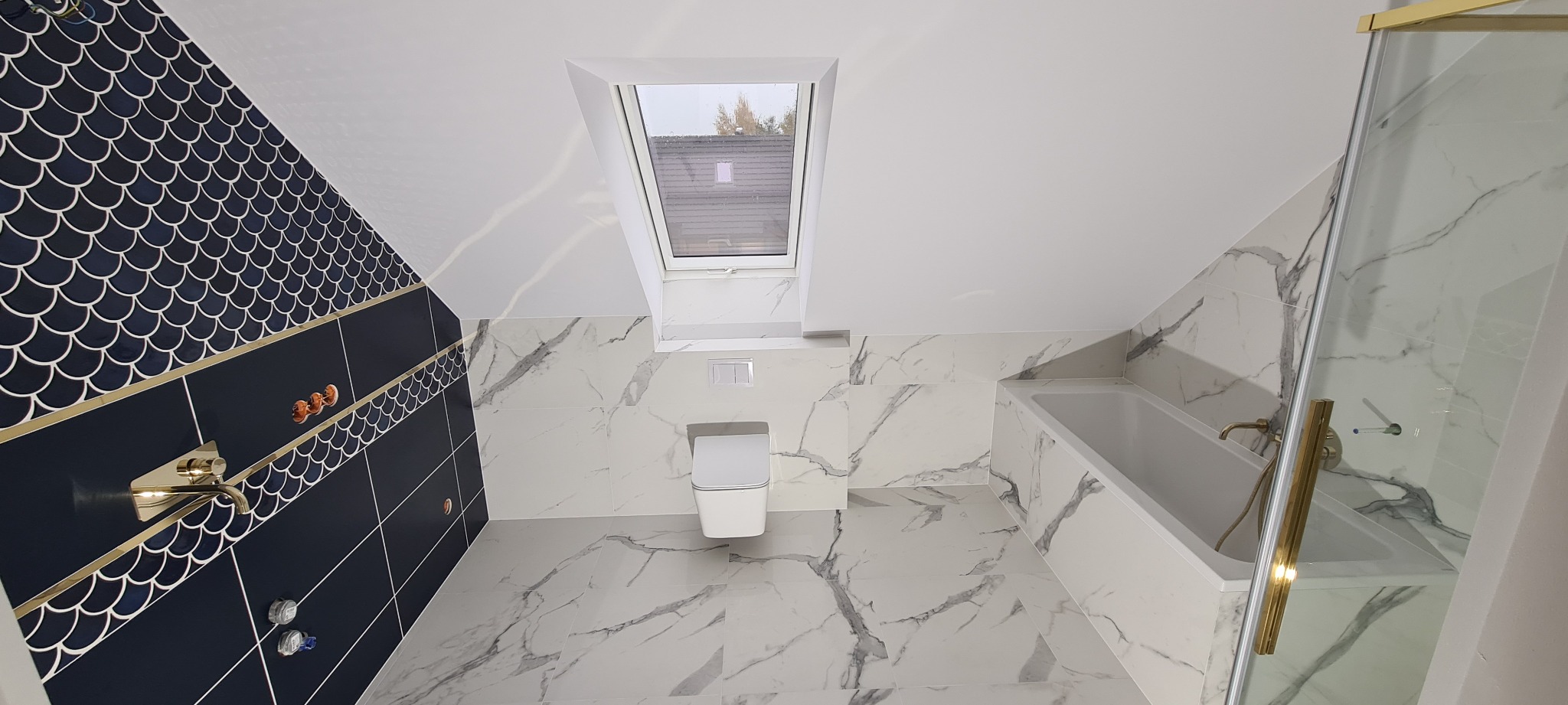 Nowoczesna łazienka na poddaszu z oknem dachowym, biało-czarne płytki imitujące marmur na podłodze i ścianach, akcenty w kolorze złotym na armaturze i kabinie prysznicowej, ściana z granatowych...