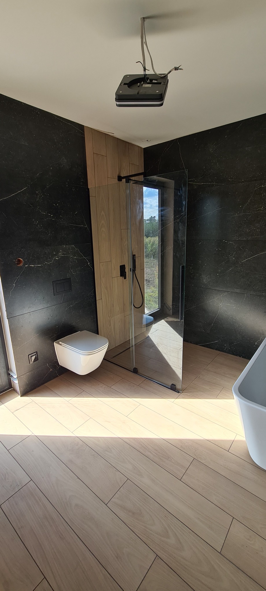 Nowoczesna łazienka z czarnymi marmurowymi ścianami, drewnopodobnymi panelami, kabiną prysznicową typu walk-in i podwieszaną toaletą; widoczna niezakończona instalacja oświetleniowa sufitowa.