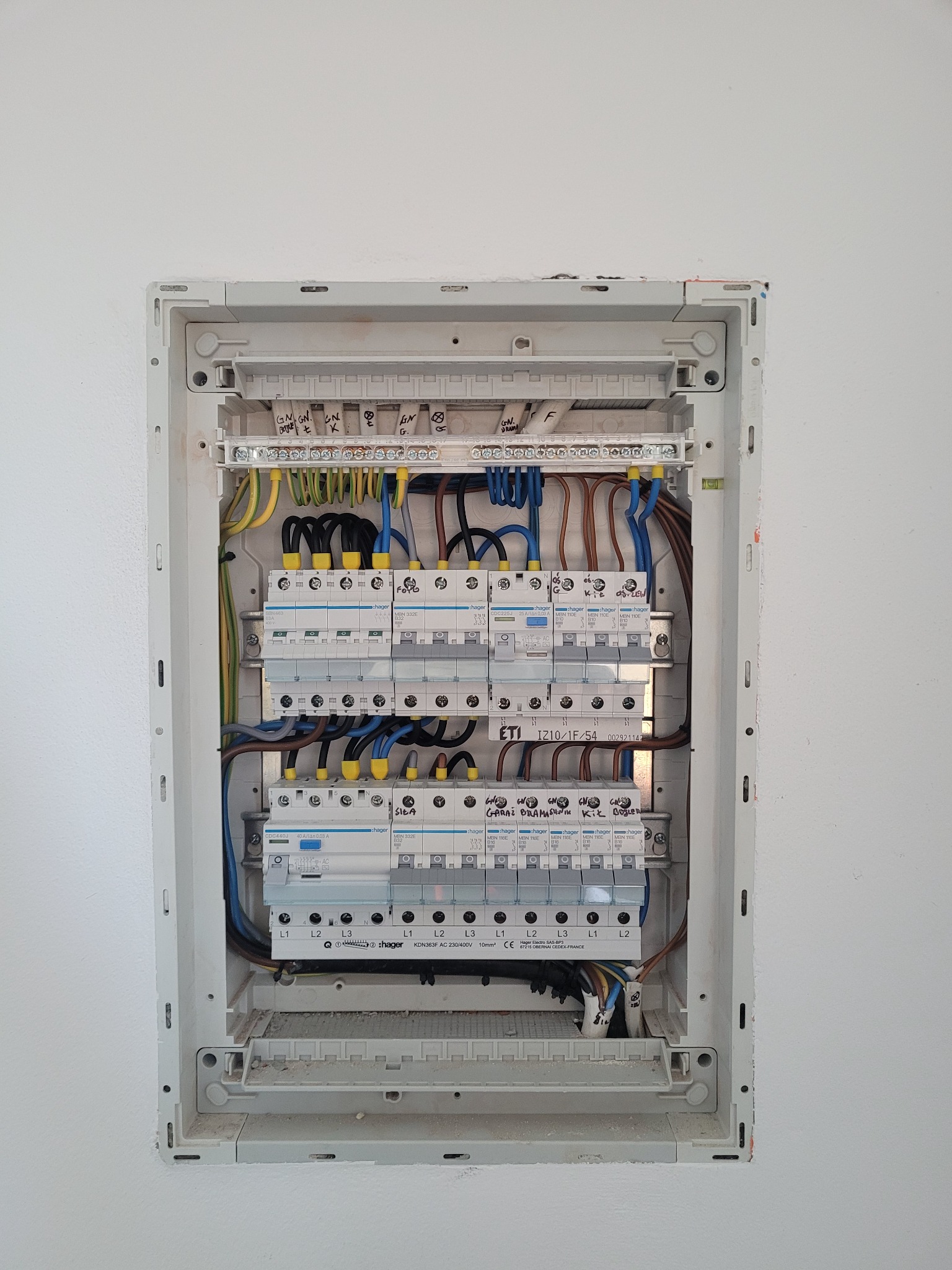 Rozdzielnica elektryczna z zainstalowanymi bezpiecznikami i okablowaniem wewnątrz obudowy, widoczne opisy markerem na listwach zaciskowych i obudowie, umieszczona na białej ścianie.