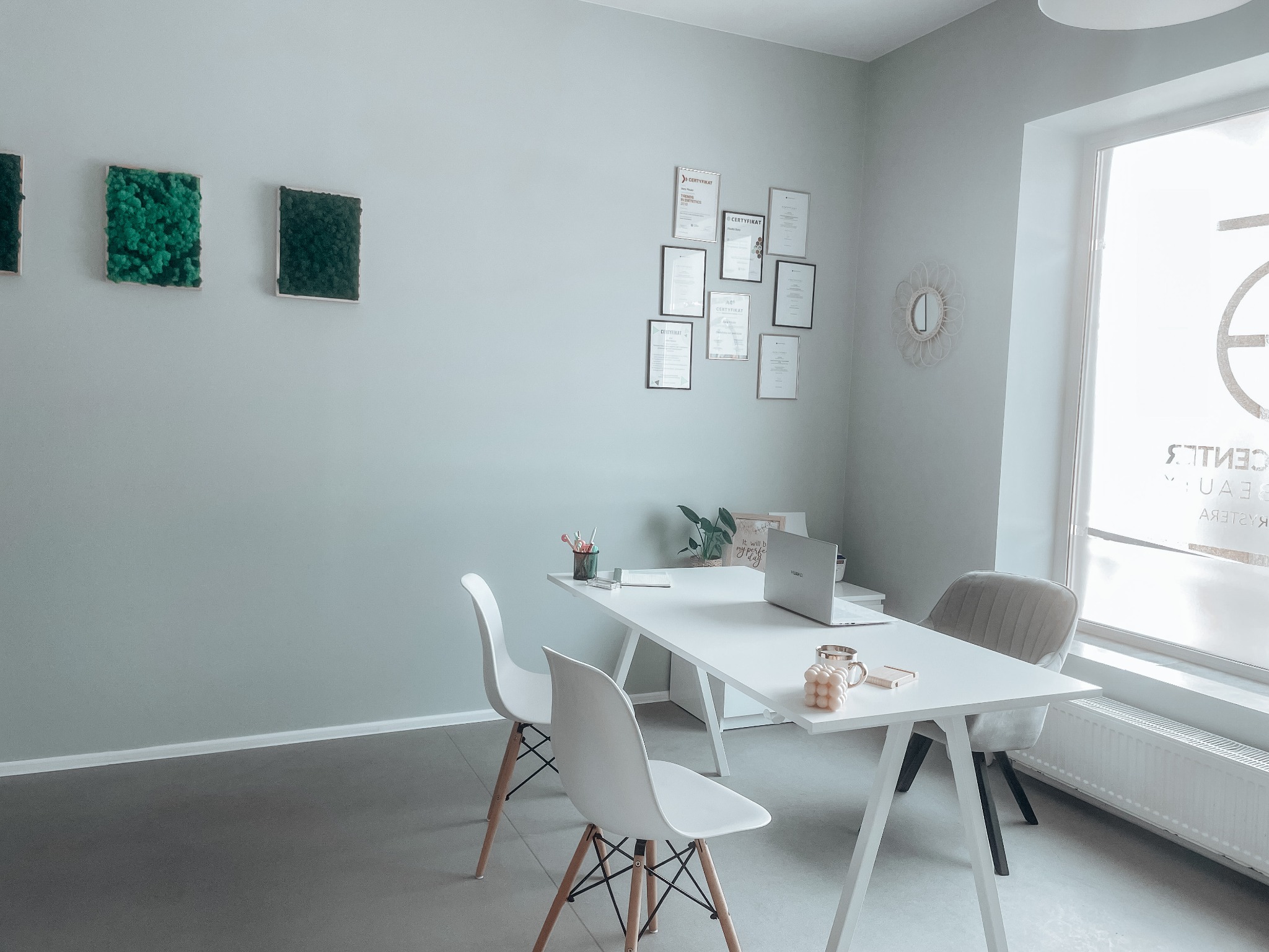 Jasne biuro z białym biurkiem, laptopem, dekoracjami i certyfikatami na ścianie, trzy zielone obrazy z mchu na ścianie po lewej stronie