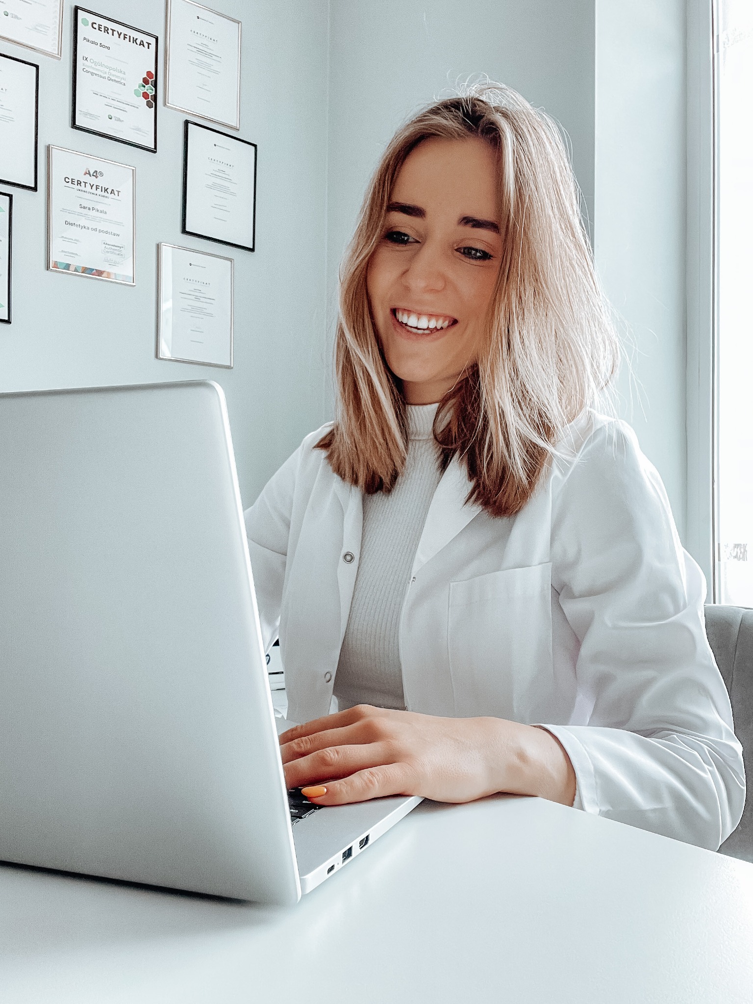 Uśmiechnięta kobieta w białym fartuchu lekarskim siedzi przy biurku z laptopem, w tle ściana z certyfikatami.