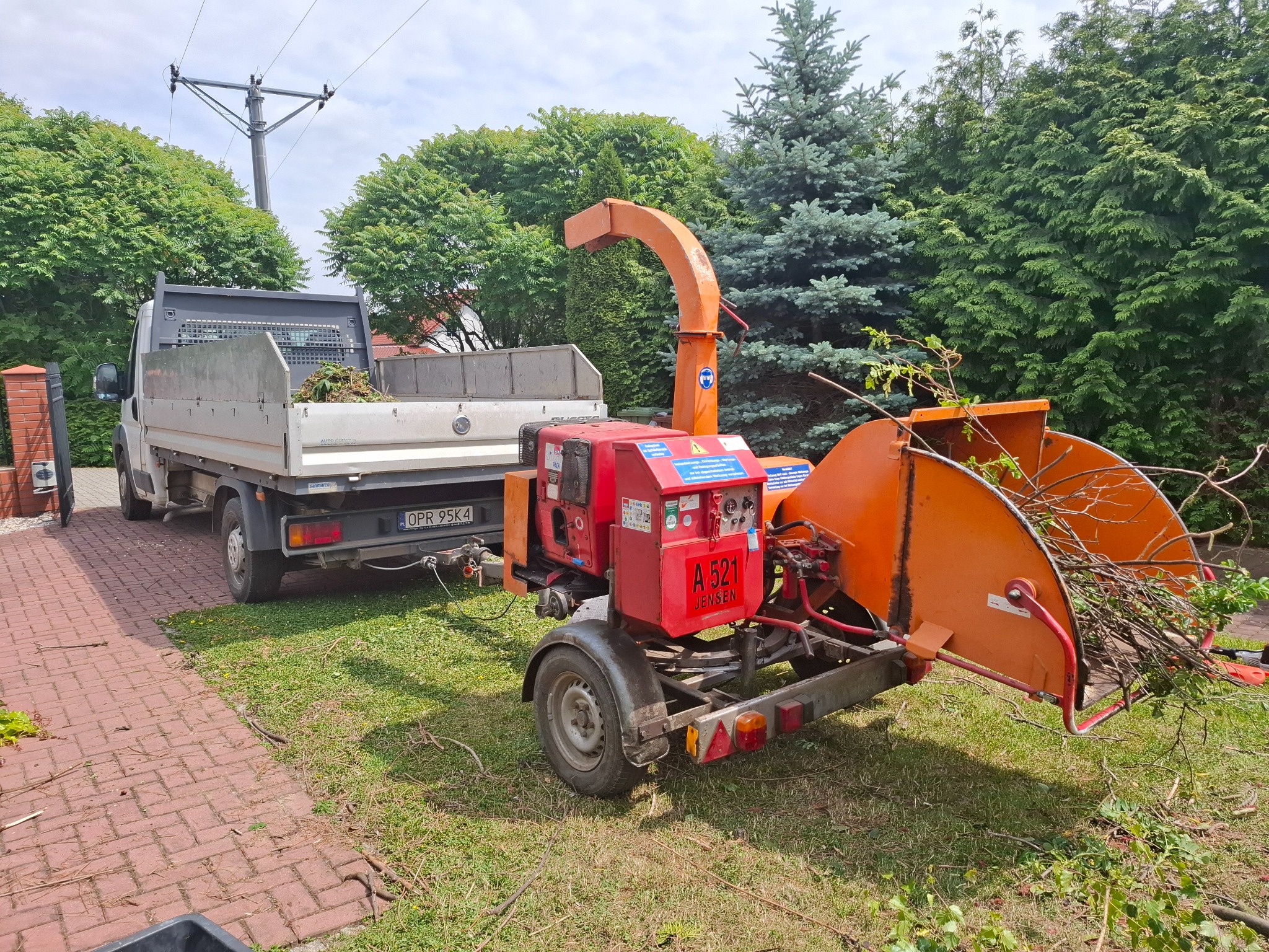 Ciężarówka z przyczepionym rębakiem do gałęzi, wypełnionym świeżo ściętymi gałęziami, stoi na trawniku przed domem.