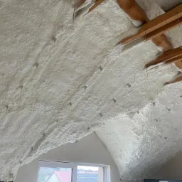 Poddasze z widoczną białą pianką izolacyjną, przymocowaną do drewnianych belek za pomocą metalowych prętów, okno w tle.