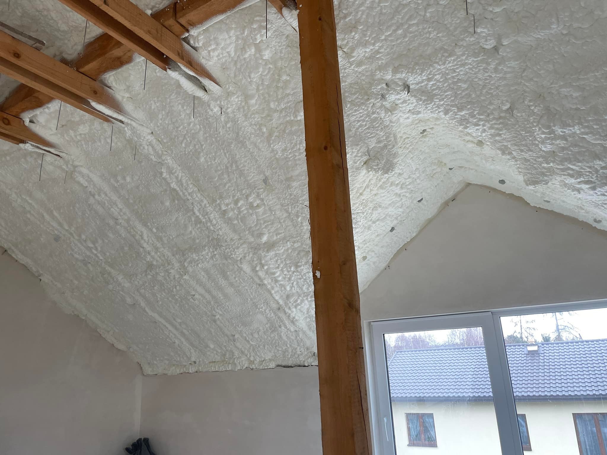 Poddasze z drewnianą konstrukcją pokryte białą pianą izolacyjną, widok na okno i dom sąsiada.