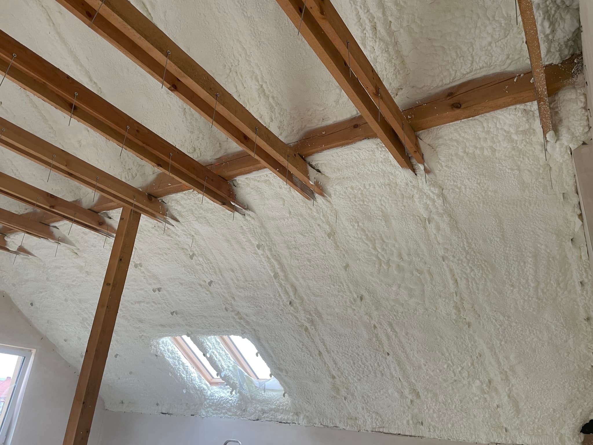 Poddasze z drewnianą konstrukcją i widoczną warstwą izolacji natryskowej, z oknami dachowymi w tle.