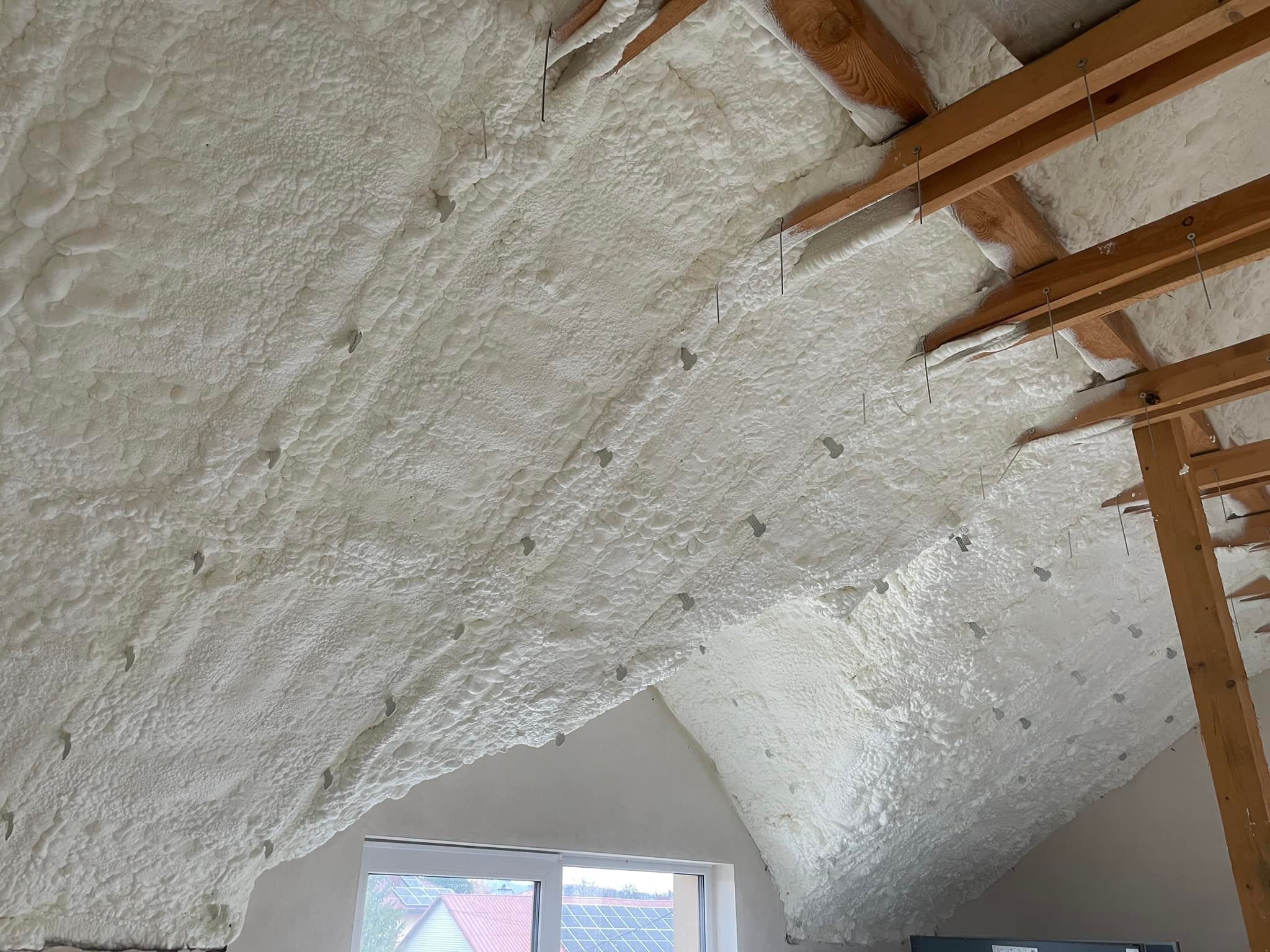 Poddasze z widoczną białą pianką izolacyjną, przymocowaną do drewnianych belek za pomocą metalowych prętów, okno w tle.