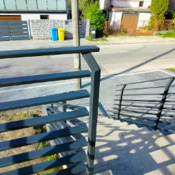 Nowoczesna, ciemnoszara balustrada metalowa z poziomymi prętami, zamontowana przy schodach zewnętrznych prowadzących do budynku mieszkalnego, widok z góry na schody i fragment ulicy z pojemnikami...