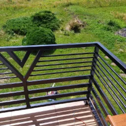 Czarna, metalowa balustrada balkonowa z poziomymi prętami i trójkątnym wzorem, widok z góry na zielony trawnik i krzewy, narzędzia w tle.