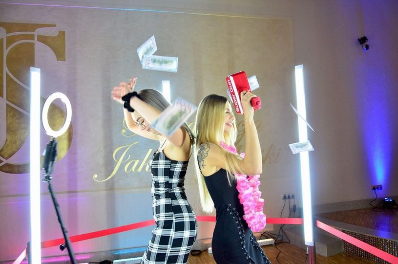 Dwie kobiety pozują tyłem do siebie, jedna z pistoletem na pieniądze, druga z unoszącymi się banknotami, na tle ściany z napisem i oświetleniem LED.