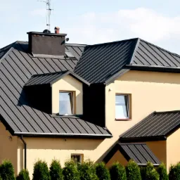 Budynek z ciemnoszarym dachem z blachy trapezowej, widoczne kominy, okna dachowe i rynny.