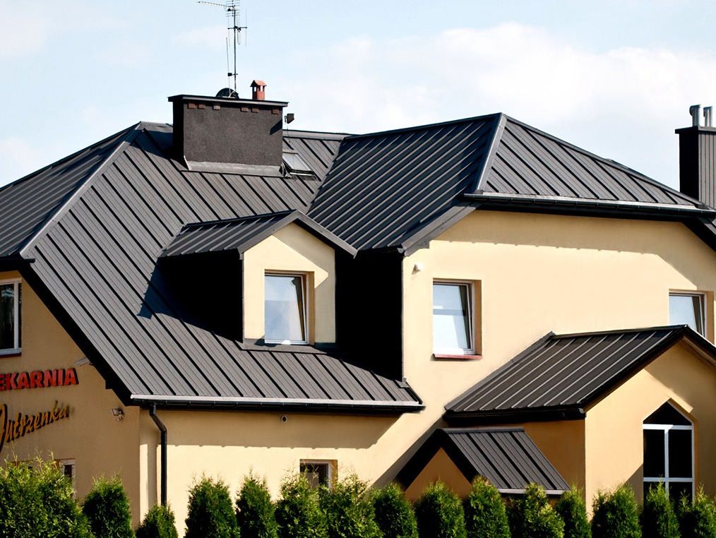 Budynek z ciemnoszarym dachem z blachy trapezowej, widoczne kominy, okna dachowe i rynny.