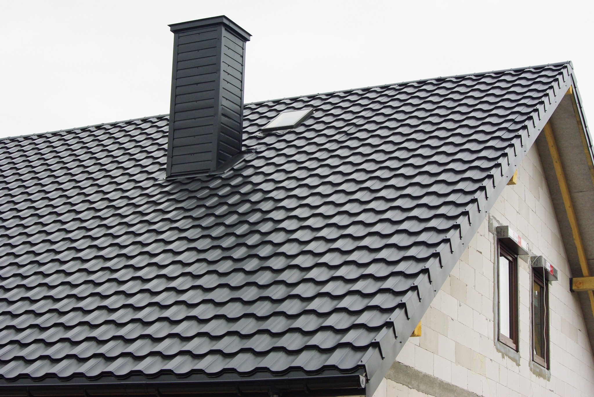 Nowy, czarny dach z blachodachówki z kominem i oknem dachowym na domu w trakcie budowy.