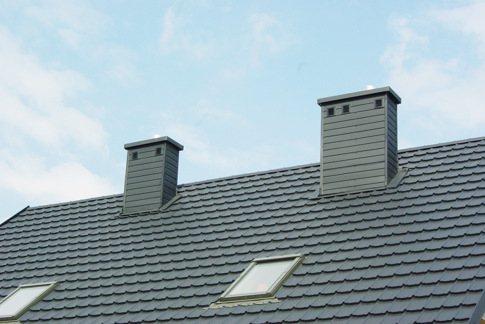 Nowoczesny dach z szarej blachodachówki modułowej, dwa kominy wentylacyjne z szarej blachy i okno dachowe, widok z dołu na tle jasnego nieba.