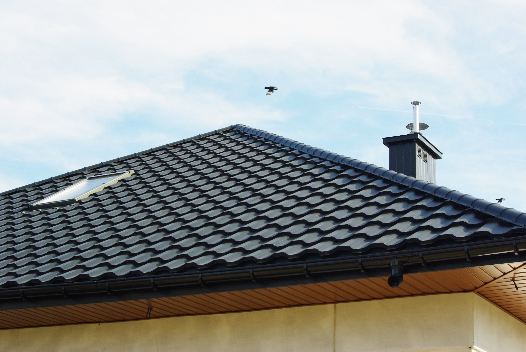 Czarny dach z blachodachówki modułowej, okno dachowe, komin z blachy i rynny, na tle błękitnego nieba z ptakami.