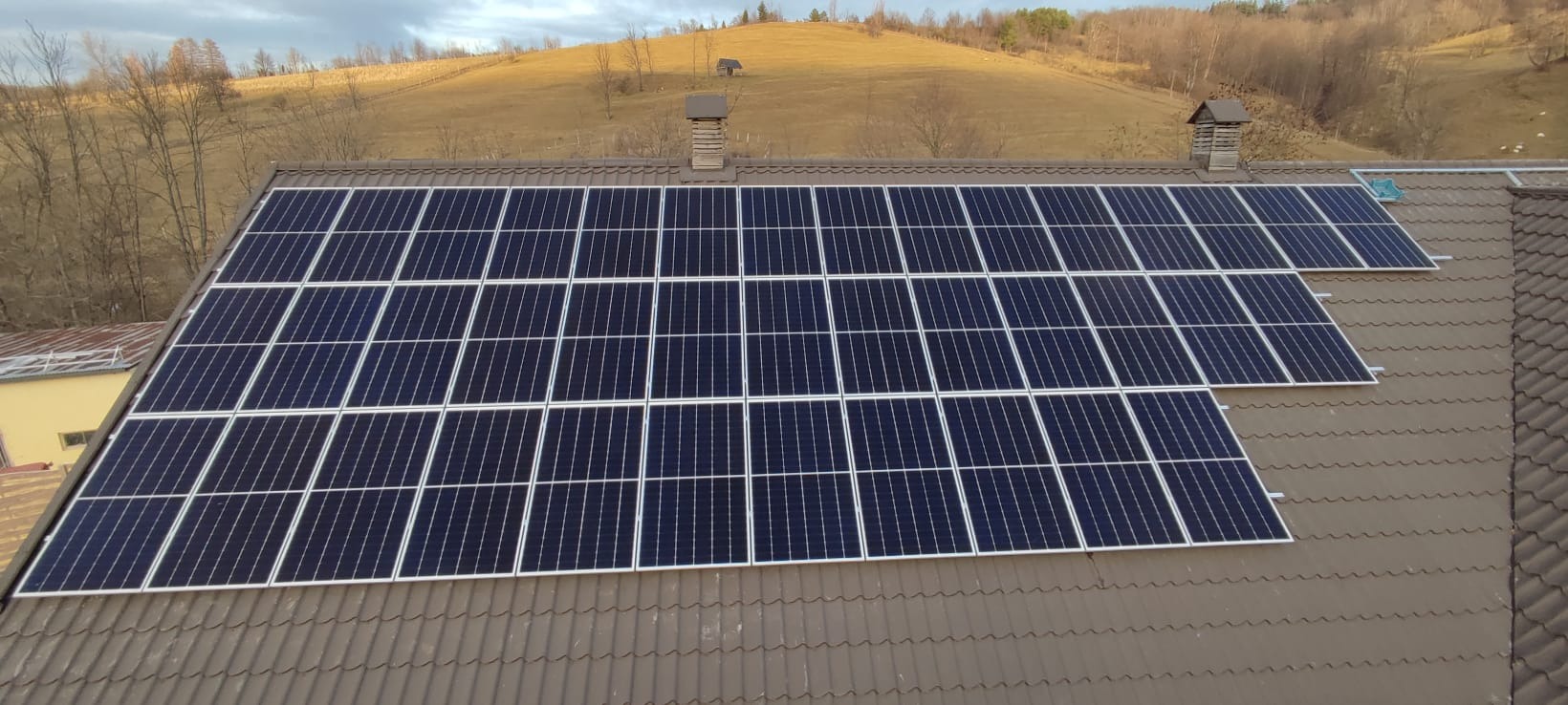 Panel słoneczny na brązowym dachu domu z widokiem na pagórkowaty krajobraz w tle.