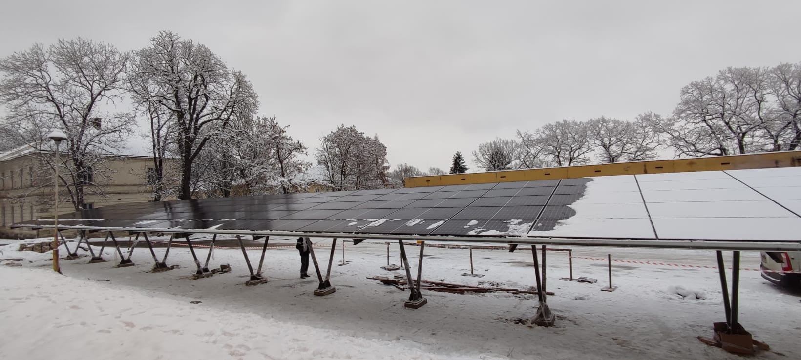 Instalacja paneli słonecznych pod śniegiem; widoczne panele na konstrukcji wsporczej, częściowo zasypane śniegiem, w tle ośnieżone drzewa i budynek.