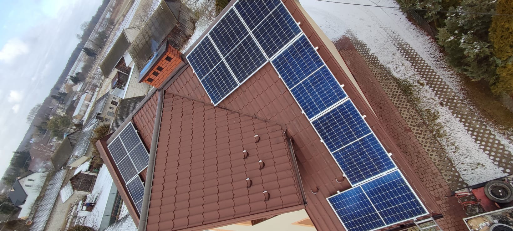 Montaż paneli fotowoltaicznych na brązowym dachu z ceglanym kominem, widok z góry na częściowo ośnieżone otoczenie budynku.