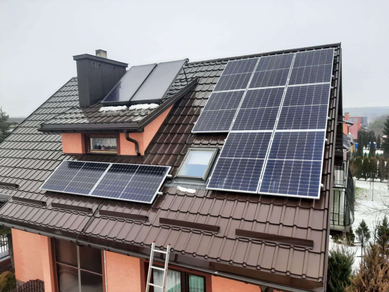 Instalacja paneli słonecznych na dachu dwuspadowym z brązowej dachówki, widoczna drabina oparta o ścianę budynku, fragmenty śniegu na dachu i w tle.