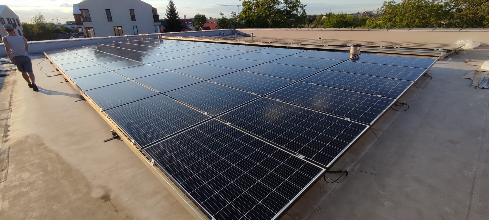 Instalacja paneli słonecznych na płaskim dachu budynku mieszkalnego, widoczny pracownik w oddali oraz elementy montażowe.