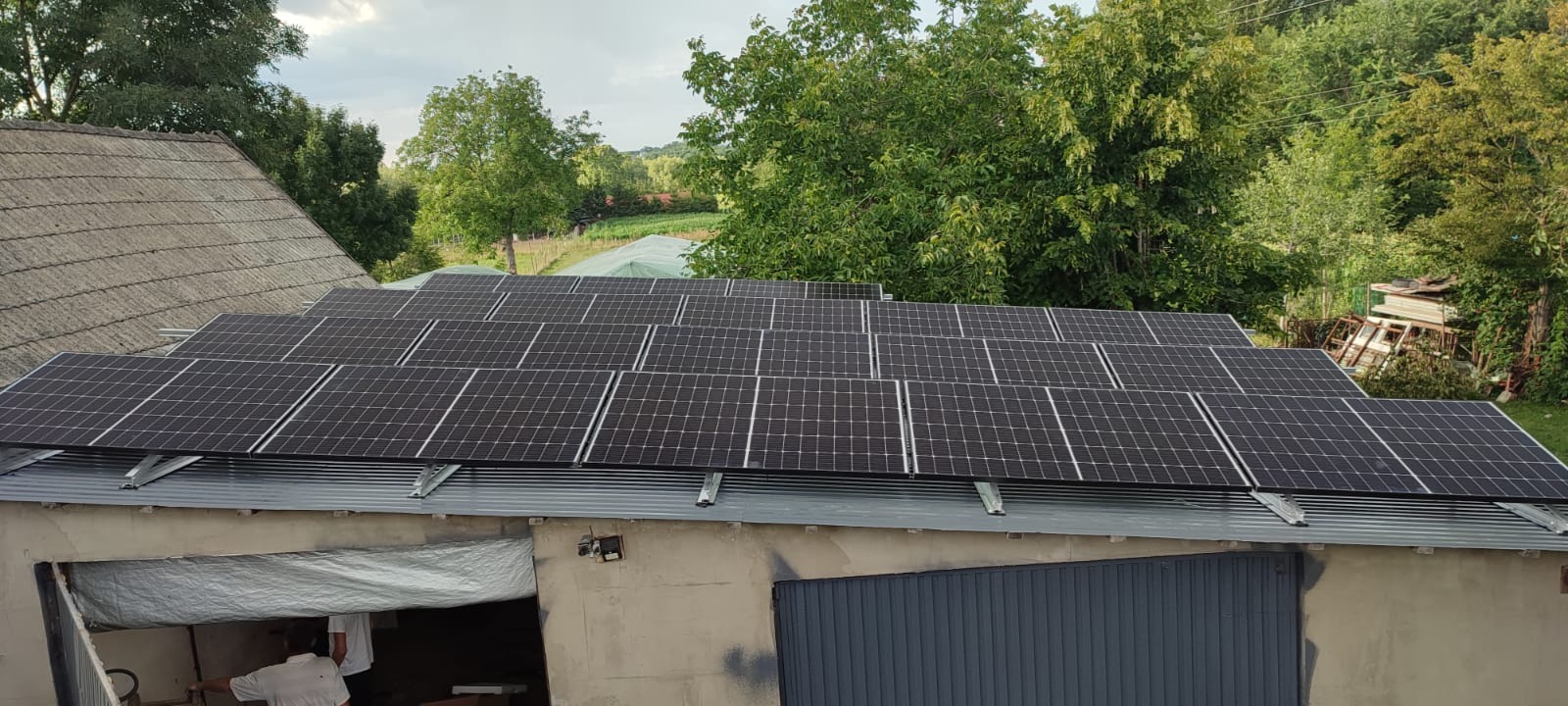 Instalacja paneli słonecznych na blaszanym dachu garażu z widocznym fragmentem starego dachu budynku obok i zielenią w tle.