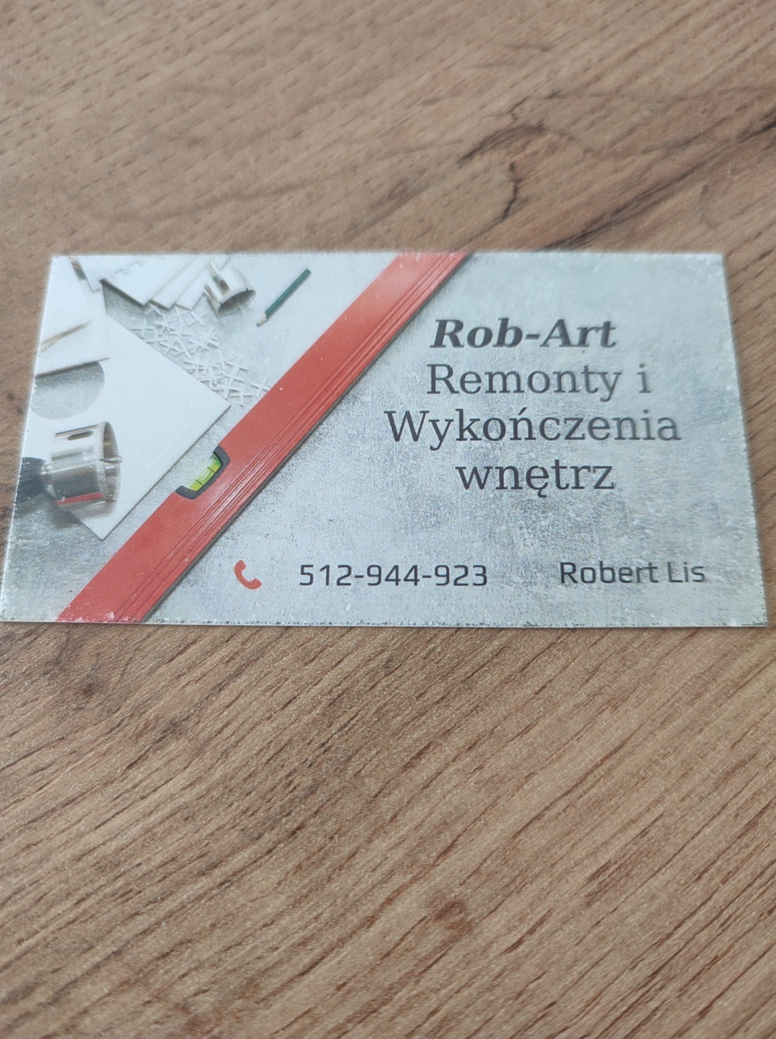 Wizytówka firmy Rob-Art Remonty i Wykończenia Wnętrz z numerem telefonu 512-944-923 i imieniem Robert Lis. Na wizytówce grafika z poziomnicą i narzędziami.