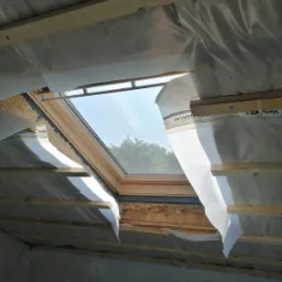 Poddasze w trakcie izolacji, widoczne wełna mineralna, folia paroizolacyjna z nadrukiem Isover Vario KB 1, drewniane krokwie i okno dachowe z widokiem na drzewa.