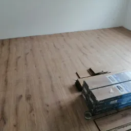 Układanie paneli podłogowych w pokoju, widoczne paczki paneli Logoclic Aqua Protect i fragment ułożonej podłogi imitującej drewno, przy ścianie widoczne niedoskonałości wymagające wykończenia.