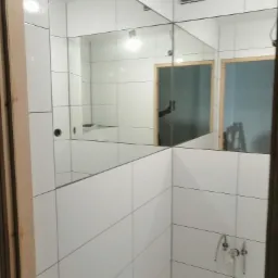 Wykończona łazienka z białymi, kwadratowymi płytkami, widoczne lustro złożone z kilku elementów, odbijające fragment pomieszczenia z drabiną malarską w tle.