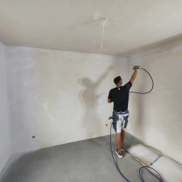 ProfibauPol - Pracownik w trakcie natryskowego wyrównywania powierzchni ścian w pomieszczeniu, widoczne narzędzia i sprzęt, ślady po szpachlowaniu na ścianach.
