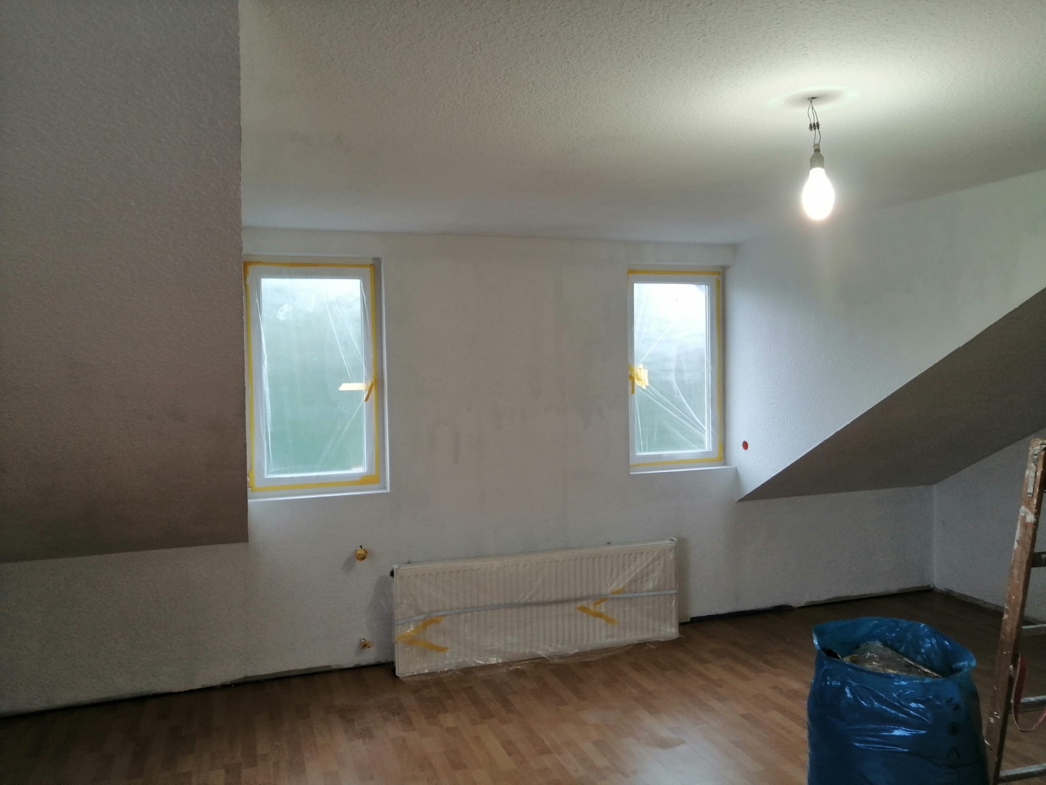 Pokój na poddaszu w trakcie przygotowań do prac wykończeniowych: okna i grzejnik zabezpieczone folią malarską, ściany po wstępnym szpachlowaniu, widoczny worek na śmieci i drabina.