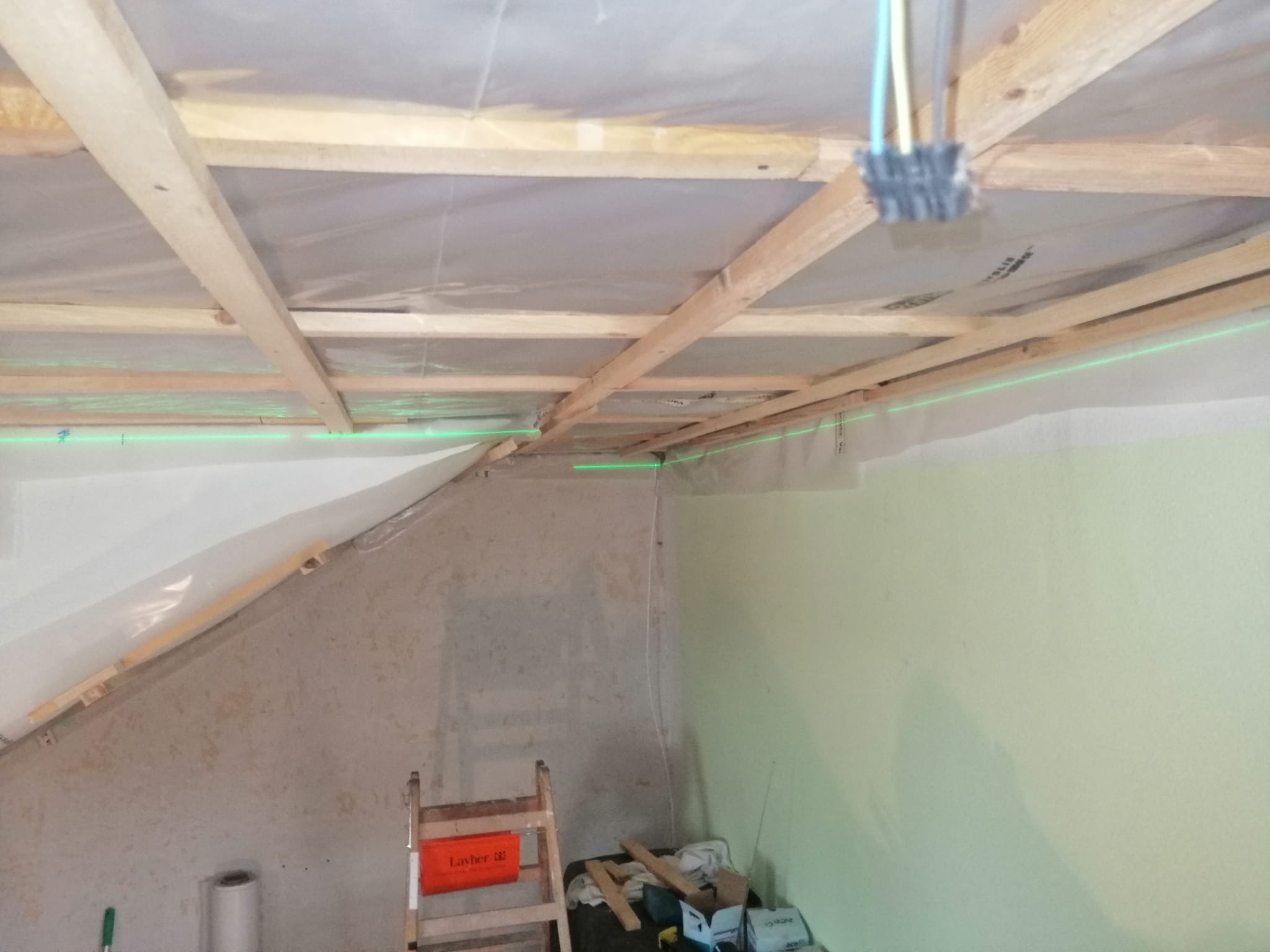 Konstrukcja drewniana pod ocieplenie poddasza z widocznymi liniami lasera wyznaczającymi poziom, drabina Layher i materiały budowlane w tle.