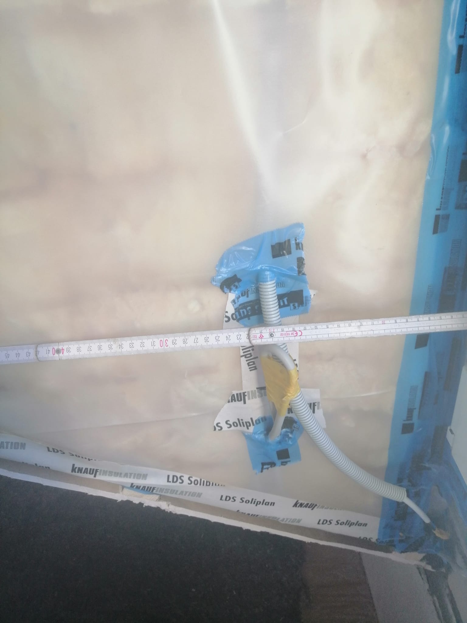 Poddasze w trakcie ocieplania, widoczna folia paroizolacyjna Knauf Insulation LDS Soliplan, peszel instalacyjny przymocowany taśmą, oraz miarka rozłożona wzdłuż izolacji.