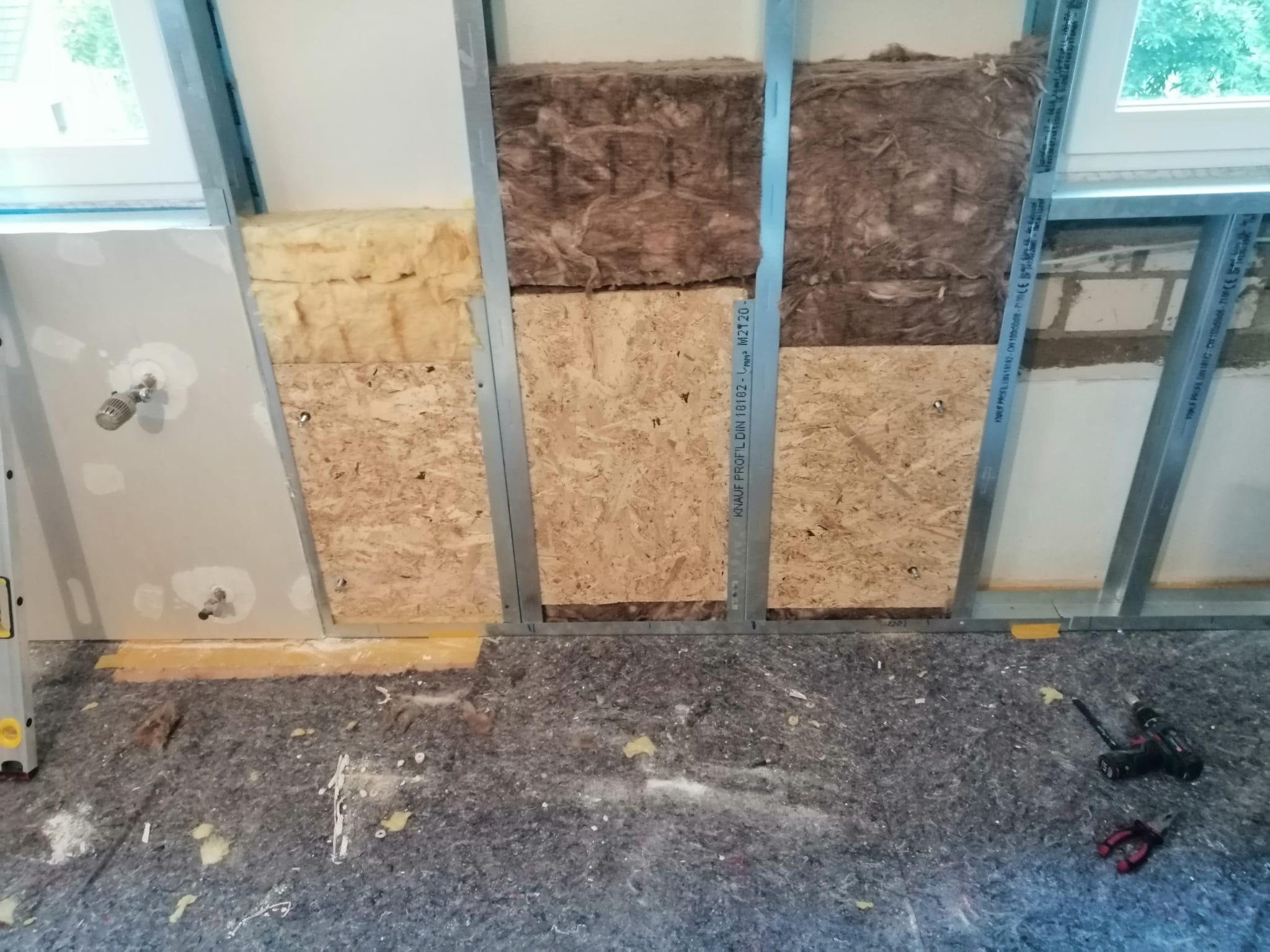 Montaż izolacji z wełny mineralnej i płyt OSB w konstrukcji szkieletowej poddasza, widoczne profile metalowe, fragment okna dachowego i narzędzia na podłodze zabezpieczonej folią malarską.