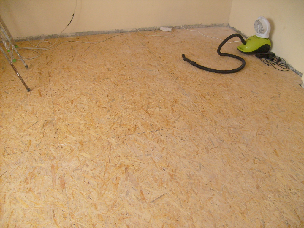 Przygotowane podłoże z płyt OSB pod przyszłą podłogę, widoczny fragment drabiny malarskiej, odkurzacz parowy i kable przy ścianie.