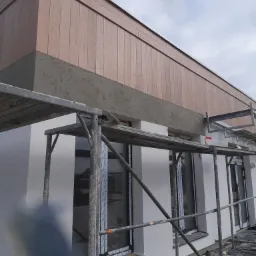 Budynek w trakcie prac elewacyjnych: widoczne rusztowanie, ocieplona ściana w kolorze białym i drewniana okładzina pod dachem, fragment czarnej folii zabezpieczającej.