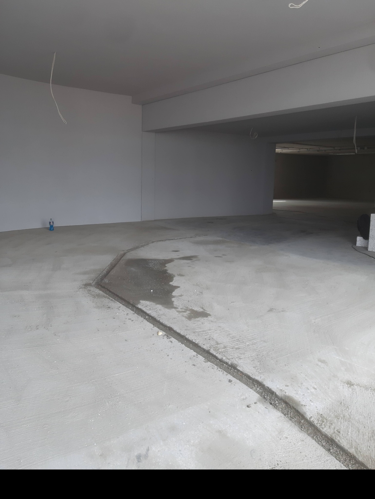 Surowy betonowy garaż z widocznymi śladami wilgoci i niedokończoną dylatacją, butelka wody w tle.
