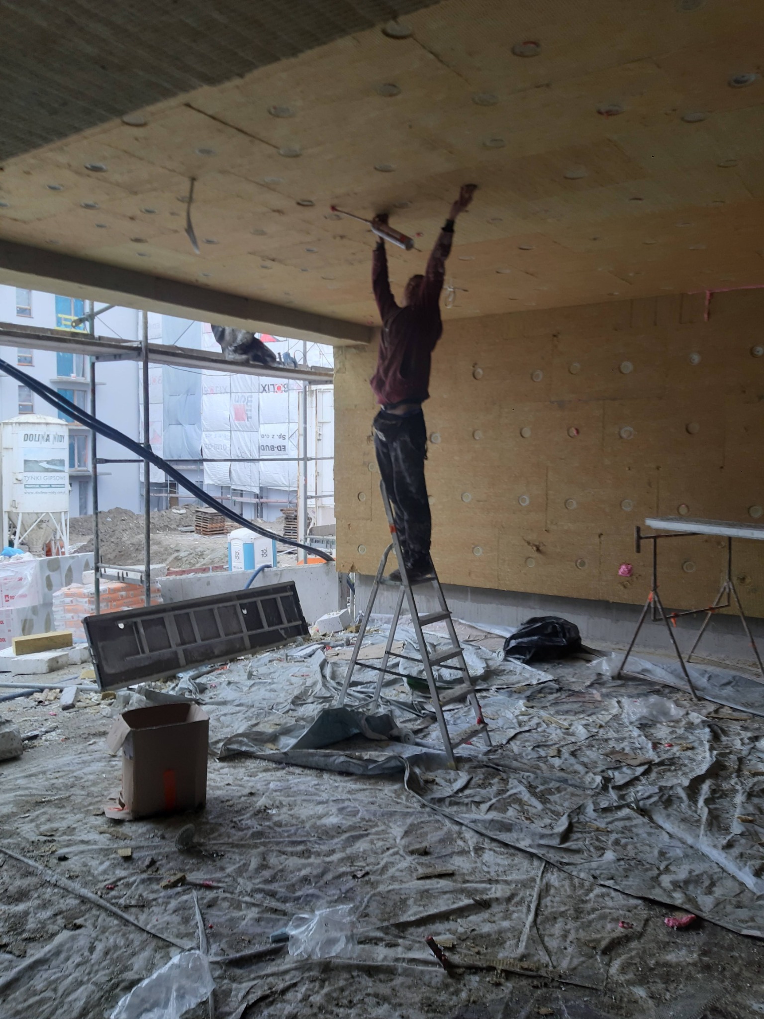 Pracownik na drabinie montuje izolację na suficie w surowym wnętrzu, widoczne płyty izolacyjne z okrągłymi otworami, zabezpieczona folią podłoga i częściowo widoczne budynki w tle.