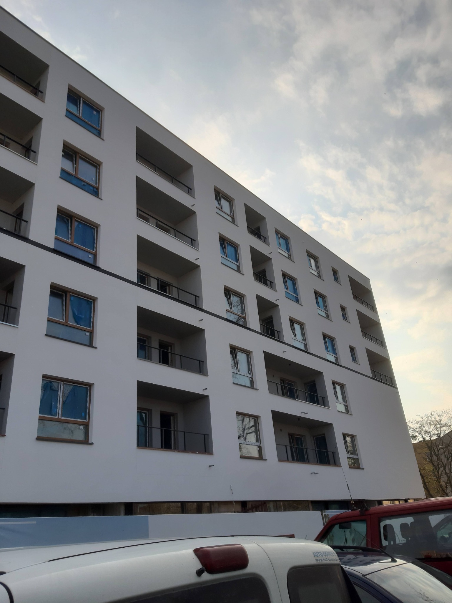 Nowy, biały budynek wielorodzinny z balkonami, okna zabezpieczone folią, widok od dołu, częściowo zasłonięty przez zaparkowane samochody.