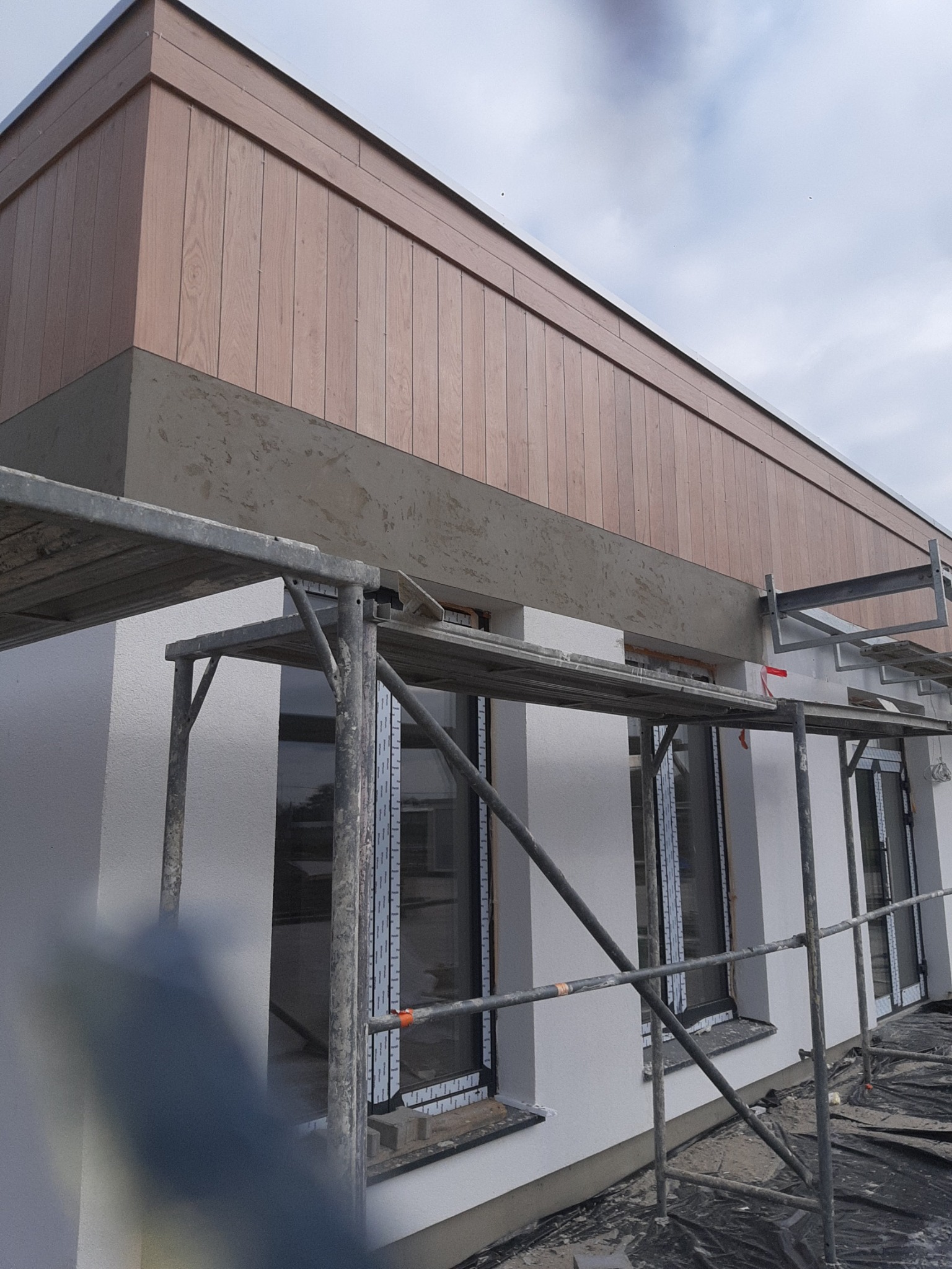 Budynek w trakcie prac elewacyjnych: widoczne rusztowanie, ocieplona ściana w kolorze białym i drewniana okładzina pod dachem, fragment czarnej folii zabezpieczającej.