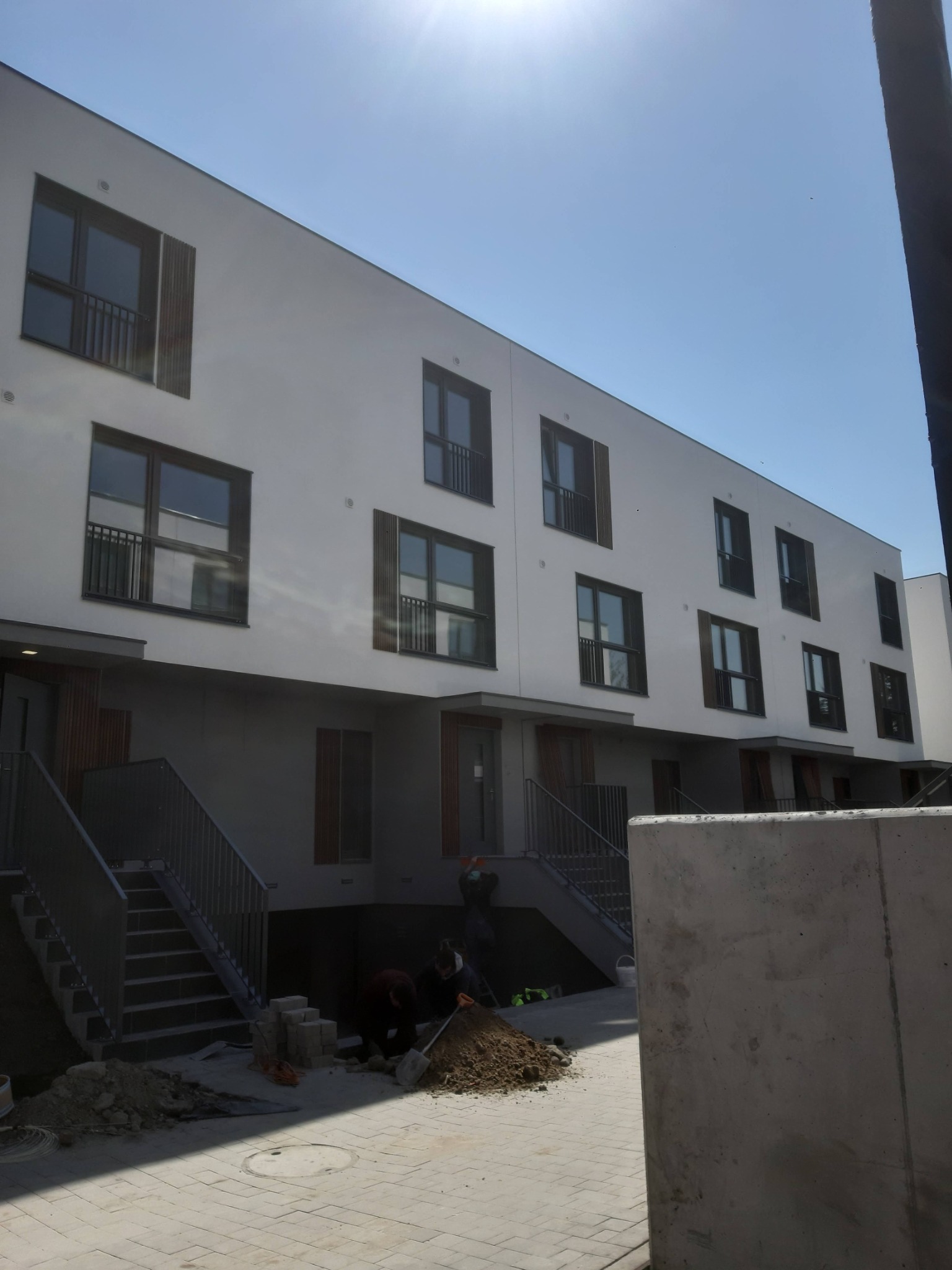 Nowoczesny budynek wielorodzinny w trakcie prac ziemnych przy wejściach, widoczne schody i pracownicy z łopatami.