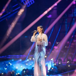 Foto 3 - Młoda kobieta w białej sukni śpiewa do mikrofonu na podświetlonej scenie z publicznością w tle.