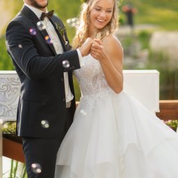 Foto 3 - Szczęśliwa para młoda trzymająca się za ręce, otoczona bańkami mydlanymi; panna młoda w koronie z zielonych liści, pan młody w ciemnym garniturze z muszką.