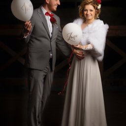 Foto 3 - Para młoda trzymająca białe balony z napisem 'Mąż' i 'Żona', mężczyzna w szarym garniturze z czerwoną muchą, kobieta w białej sukni i futrzanym etolu, czerwone róże we włosach i butonierce.