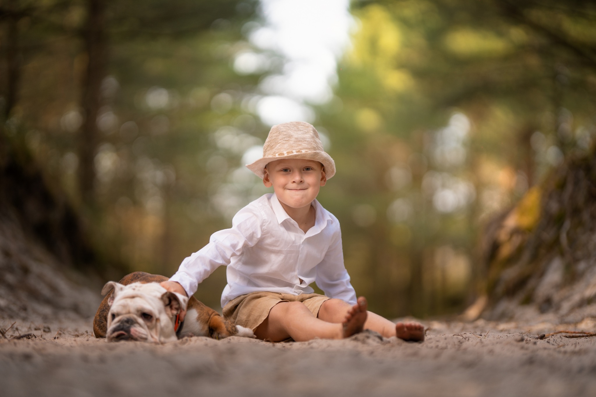 Chłopiec w słomkowym kapeluszu i białej koszuli siedzi na piaszczystej ścieżce w lesie, trzymając rękę na leżącym obok buldogu angielskim.