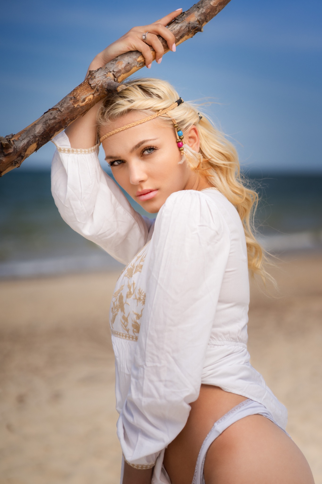 Portret blondynki w białej bluzce z haftem, opierającej się o gałąź na plaży, z dekoracyjną opaską na głowie i kolczykami z kolorowymi koralikami.