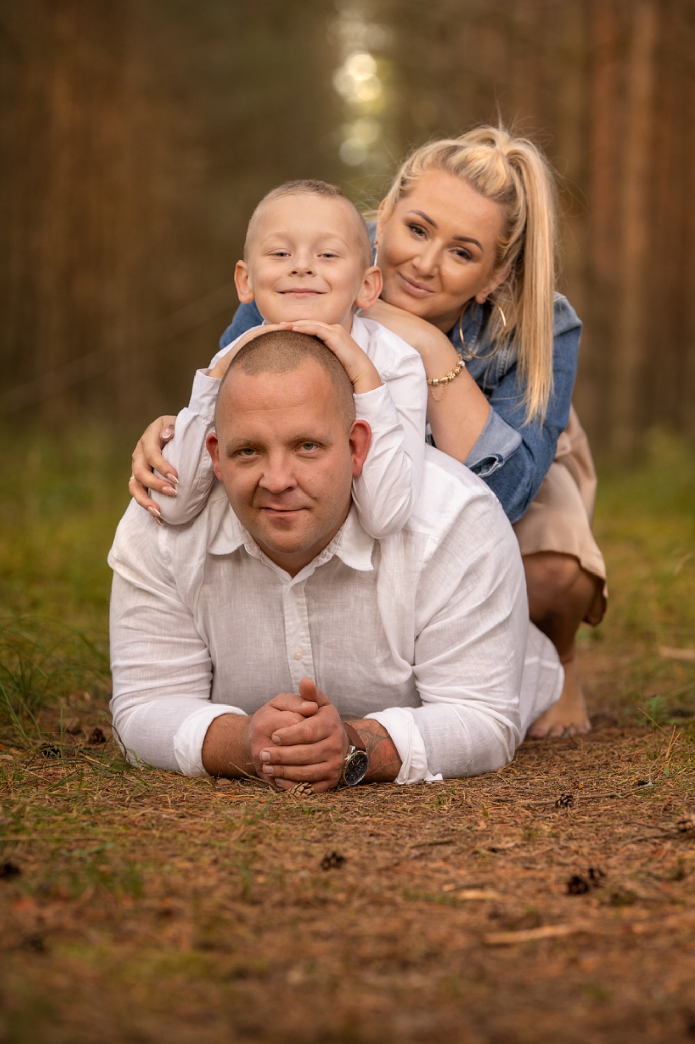 Uśmiechnięta rodzina: ojciec leży na ziemi, syn opiera się o jego głowę, matka obejmuje ich, las w tle.