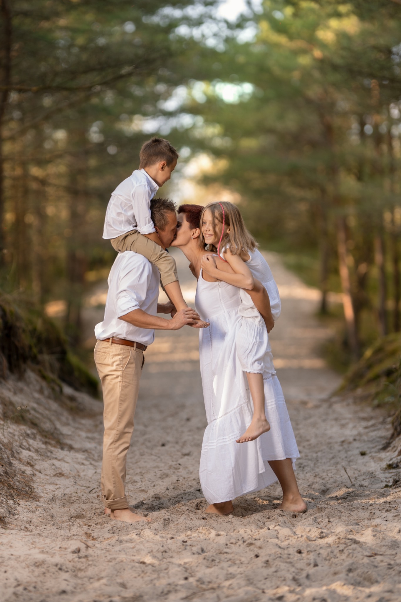 Rodzinne ujęcie w lesie: ojciec z synem na ramionach całuje matkę z córką na plecach, wszyscy boso na piaszczystej ścieżce, w bieli.