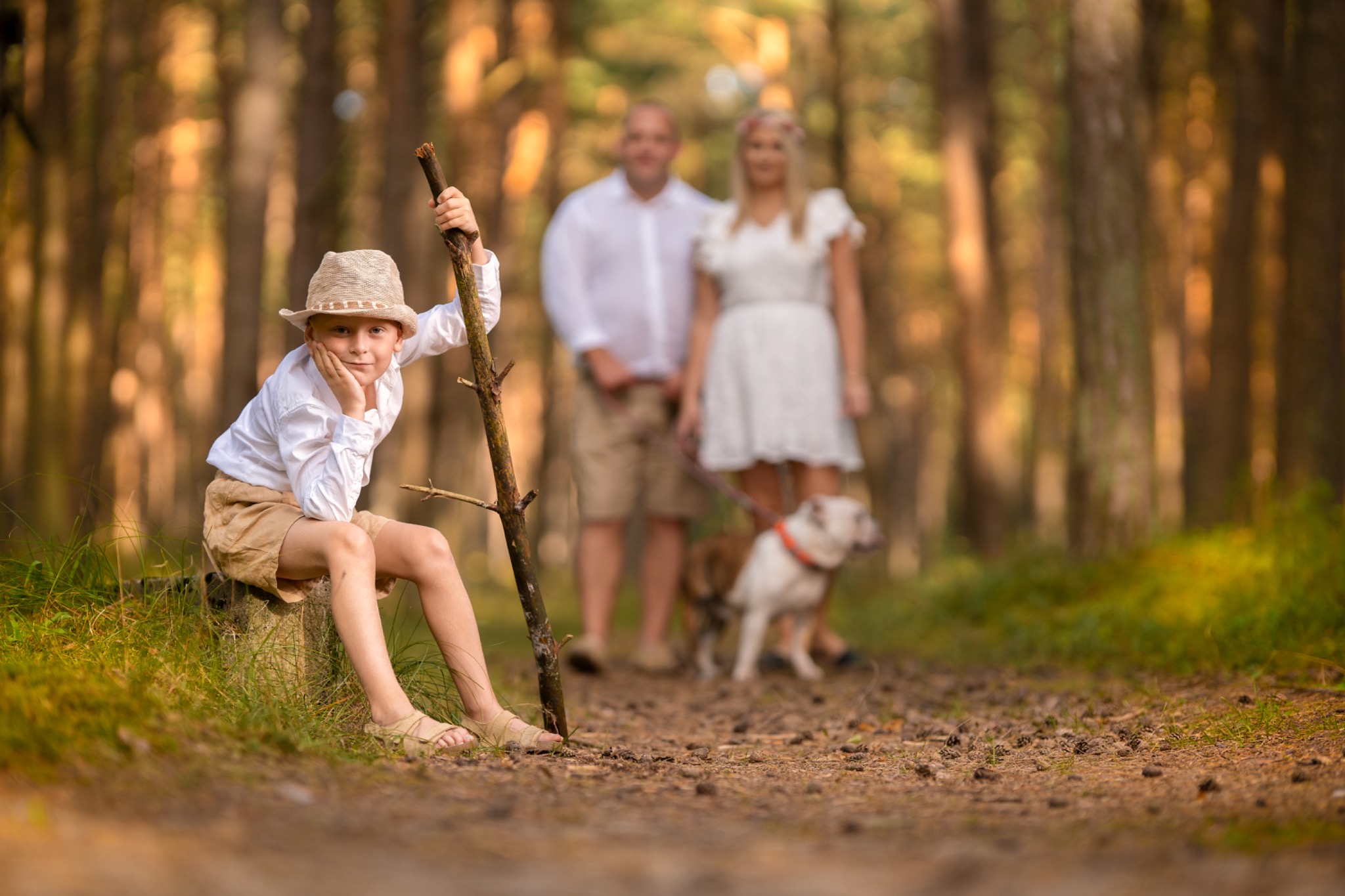 Chłopiec w słomkowym kapeluszu oparty o kij siedzi na pniu drzewa w lesie, w tle para z psami na smyczy, jesienna kolorystyka.
