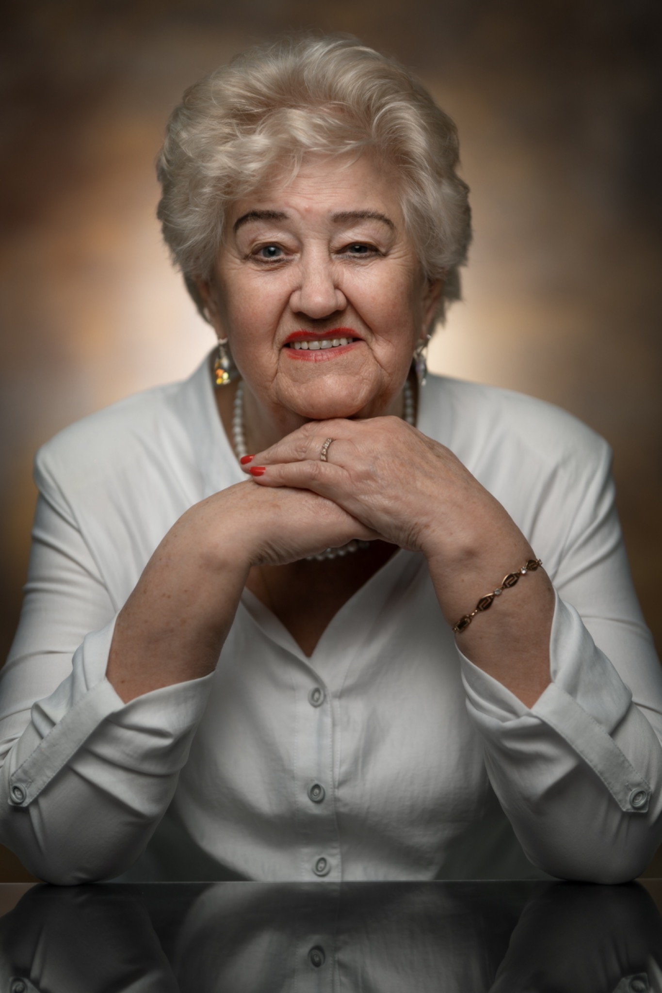 Portret starszej kobiety z platynowymi włosami, ubranej w białą koszulę, z rękami splecionymi pod brodą, na rozmytym brązowym tle.